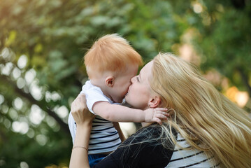 Mom and son are playing together outdoors in the summer. Mother is holding the kid boy and kissing him
