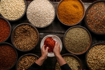 Top view of hands holding red spice powder surrounded by assorted grains and seeds in bowls on a rustic wooden table