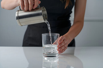 Person pouring fresh drinking water from a stainless steel pitcher into a clear glass on a white table