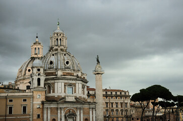 Obraz premium View of baroque church with twin domes and ancient Trajans Column against dramatic overcast sky with stone pine trees in historic Rome center