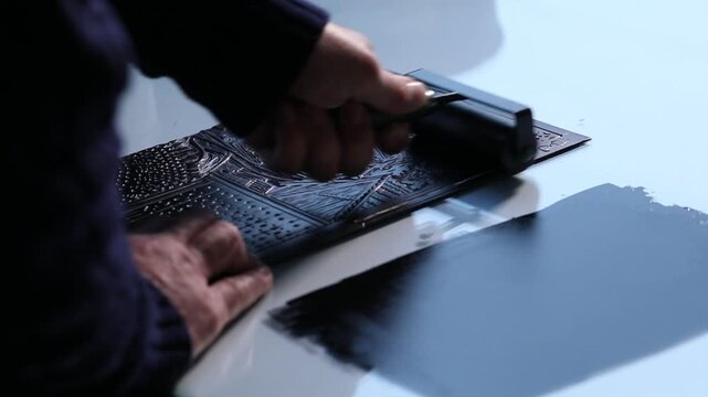Close-up of artist hands using a brayer to roll out black ink and apply it to a carved linoleum block for relief printing. Traditional art creation process.

