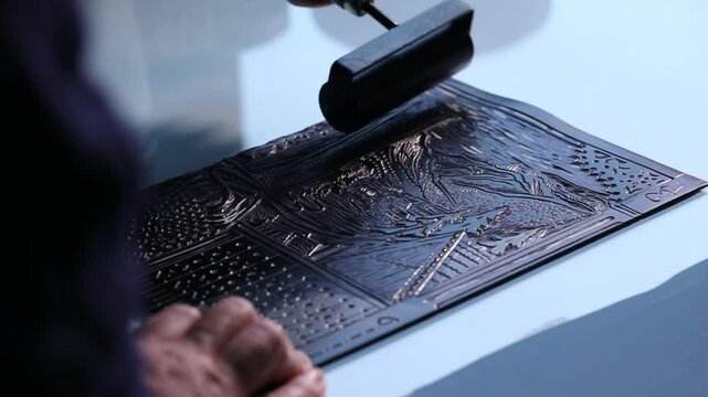 Close-up of artist hands using a brayer to roll out black ink and apply it to a carved linoleum block for relief printing. Traditional art creation process.

