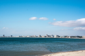The scene shows a Bulgarian sunny beach with distant buildings lining the shore. The black sea is calm and clear under a blue sky. This location attracts tourists looking to enjoy the beach