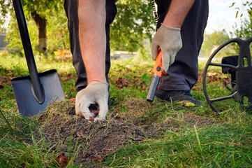 A person is crouched down in grass, using a shovel to dig in the ground. They are examining the soil with a detecting tool. Trees are nearby under a clear sky