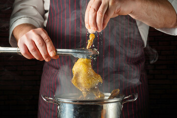 A chef uses tongs to lift a chicken leg from a pot while sprinkling salt on it. Steam rises in a busy kitchen setting during the cooking process