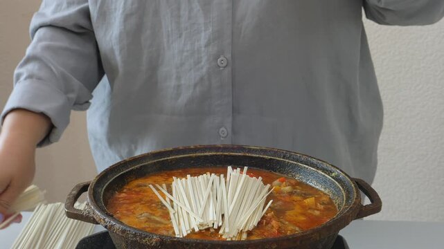 Vegetables and meat are stewed in a deep cast-iron skillet with sauce and spices, and a woman is adding udon. Medium shot.