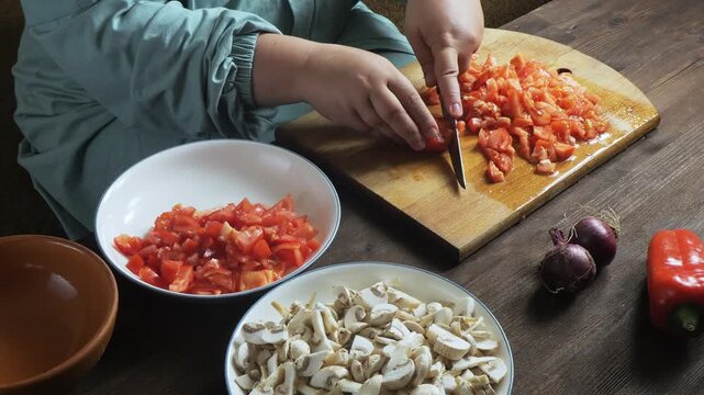 A woman's hands slice red tomatoes with a knife on a cutting board on the table and place them on a plate. Medium shot.