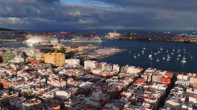 Aerial view shows Las Palmas waterfront, Puerto de la Luz, marina breakwaters, port cranes, cruise ships, sailboats, beach, promenade, and Mount Teide under heavy clouds.