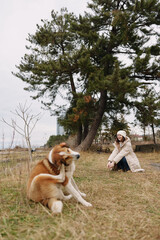 Woman in a coat and beanie sits on a grassy park, a relaxed dog nearby under a large tree, portraying calm outdoor companionship and gentle nature moments.