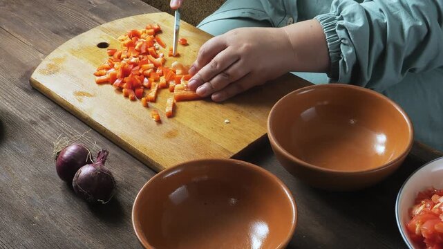 Red peppers are sliced ​​with a knife on a cutting board on the table and placed on a plate. Medium shot.
