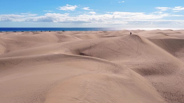 Aerial drone motion shows Maspalomas dunes on Gran Canaria, wind etched ridges, footprints, walkers, a small green oasis, and the Atlantic under scattered clouds.