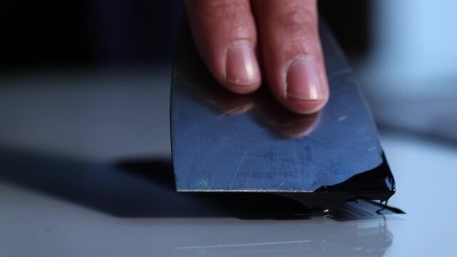 A close-up shot of an artist's hand using a palette knife to spread smooth black ink on a glass surface, preparing for a printmaking or linocut project.

