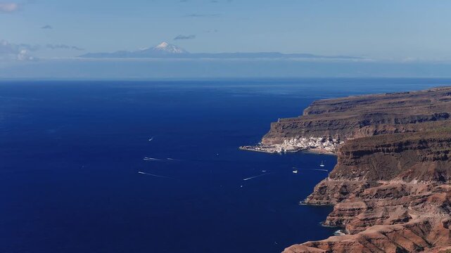 Aerial view shows La Playa de Mogan on Gran Canaria, boats leaving wakes, basalt cliffs and terraces, deep blue Atlantic, clear daylight, Mount Teide on horizon.