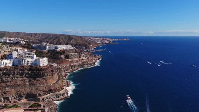 Aerial 4K view of Gran Canaria's southwest coast with RIU hotel, winding road, ochre cliffs, turquoise surf, boats, and distant Tenerife with Mount Teide.