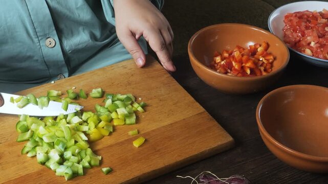Hands chop green peppers with a knife on a cutting board on the table and place them on a plate. Medium shot.