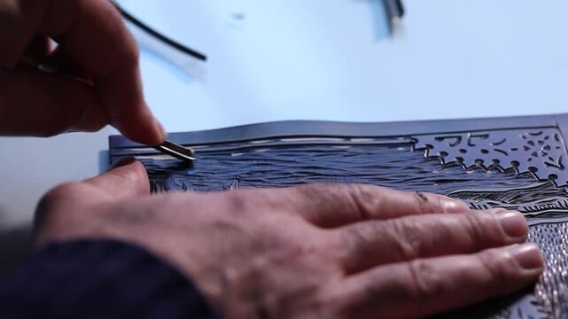 Macro shot of an artist's hands using a gouge to carve intricate details into a linoleum block, creating a relief printmaking matrix in a creative art studio.