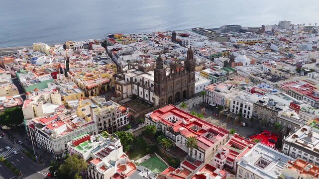 Aerial daylight pan over Vegueta, Las Palmas, Canary Islands, showing Catedral de Santa Ana, twin towers, rose window, palm plaza, red carpet, rooftops, and sea horizon.