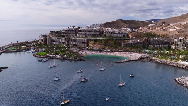 Aerial panorama shows Amadores bay in southern Gran Canaria, a sheltered beach with rock breakwaters, marina, resorts on cliffs, arid hills, and boats drifting in calm water.
