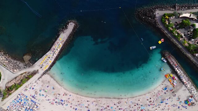 Aerial top down view of a crescent beach in Gran Canaria with stone breakwaters, yellow sunbeds, promenades, umbrellas, sunbathers, swimmers, boats, and wake trails.