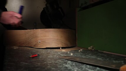 Professional luthier working on an acoustic guitar body. Close-up of male hands sanding and polishing the wooden surface in a dimly lit workshop during the manufacturing process.

