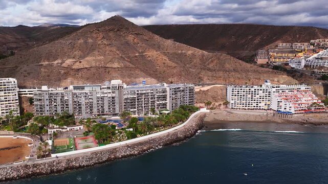 Aerial view of Gran Canaria south coast resorts, Radisson Blu sign, palm promenades, pools and courts by a sandy bay, boats, seawall, arid volcanic hills, partly cloudy light