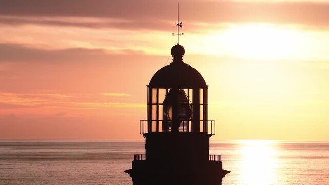 Aerial view of Faro de Maspalomas, Gran Canaria at sunset. Lantern room, Fresnel lens and weather vane silhouette. Calm sea glows, small boat drifts on horizon.