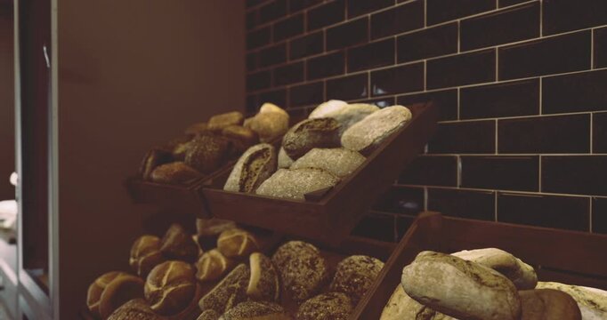 Various types of artisan bread are beautifully arranged on wooden racks in a modern bakery. The black tiled wall adds a stylish touch to the warm, inviting environment.