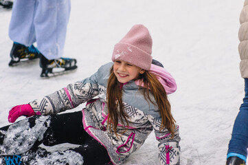 A beautiful happy teenage girl, child, lies in the snow on a snowy ice rink after slipping and falling outdoors in winter. Photography, portrait, sports, and leisure concept.