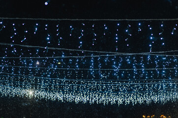 Photograph of hanging blue glowing lights, garlands against snow at night on a street in the city.