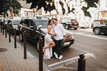 Wedding photography, close-up portrait of happy fashionable newlyweds, young stylish groom and beautiful brunette bride standing near a black car on the street in the city.