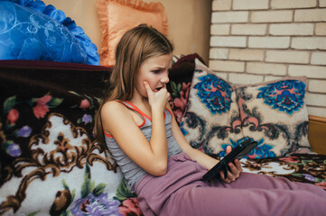 A beautiful girl, a teenage child, lies on a bed at home with a smartphone in her hands, watching a scary video, playing on a gadget, experiencing horror.