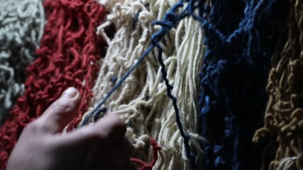 Close-up of artisan hands arranging colorful wool threads on a vertical loom. A woman is working on a handmade tapestry, carefully handling the soft red and green yarn fibers.

