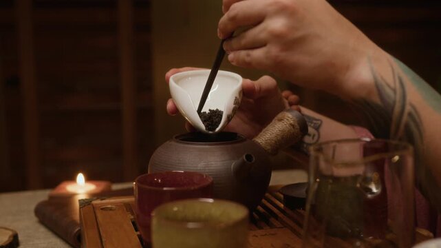 Close-up of hands of anonymous female Chinese gongfu ceremony master transferring dry tea leaves from cup into teapot with wooden spoon before brewing at home or for guests