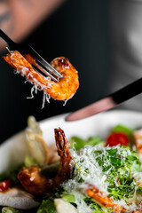 A close-up shot of a fork lifting a cooked, seasoned prawn topped with grated parmesan cheese from a fresh green salad.