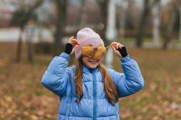 A beautiful, smiling, happy teenage girl closes her eyes with maple leaves during autumn leaf fall. Close-up portrait.
