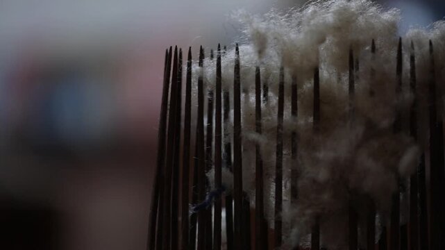 Extreme close-up of a metal weaving comb with wool fibers. The focus shifts to show a colorful, hand-woven carpet pattern on a traditional loom in the background.

