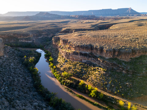 Aerial views capture the winding Virgin River as it carves through deep red sandstone canyons near Hurricane, Utah. The dramatic landscape features the massive Hurricane Cliffs and distant, majestic p