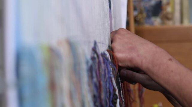 Close-up top view of artist hands weaving colorful tapestry on a loom. The artisan meticulously knots threads to create unique handmade textile wall art design.

