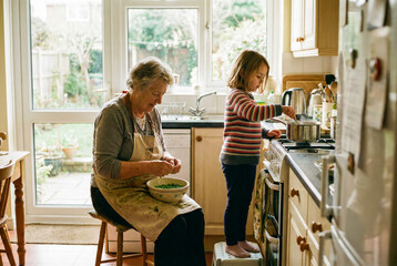 Grandmother shells peas while granddaughter stirs pot on gas cooker in bright kitchen with garden view.