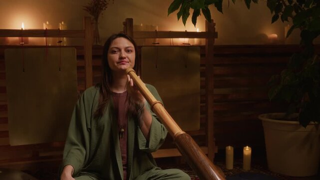 Medium portrait of Caucasian female sound healing practitioner sitting with crossed legs in lotus posture on carpet, playing didgeridoo flute, looking at camera, smiling with inner peace and happiness