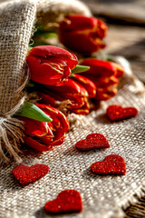 Red tulips wrapped in burlap sack lie on rustic wooden surface with glitter hearts.