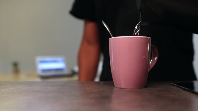 Unrecognizable person pouring boiling water from a kettle into a pink ceramic cup with a spoon inside, preparing a hot beverage at home