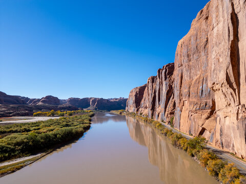 Aerial drone view of Wall Street's red sandstone cliffs in Moab at sunrise. The Colorado River winds alongside Potash Road as golden light hits climbers on the vertical walls and dark desert shadows