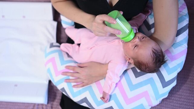 Attentive mother feeding her hungry newborn baby with a bottle of milk formula while the infant lies on a nursing pillow. Caring mom gives food to her adorable little daughter at home