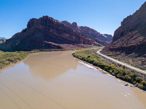 Aerial drone view of Wall Street's red sandstone cliffs in Moab at sunrise. The Colorado River winds alongside Potash Road as golden light hits climbers on the vertical walls and dark desert shadows