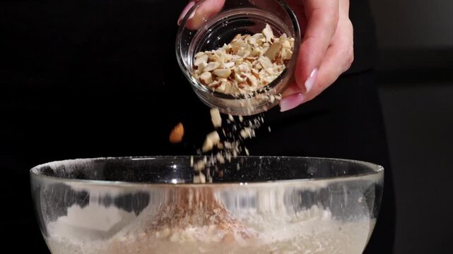 Adding chopped nuts and oats to a bowl of flour, mixing ingredients together for a healthy snack. Hand adds mixed chopped nuts into batter for banna bread or other sweets