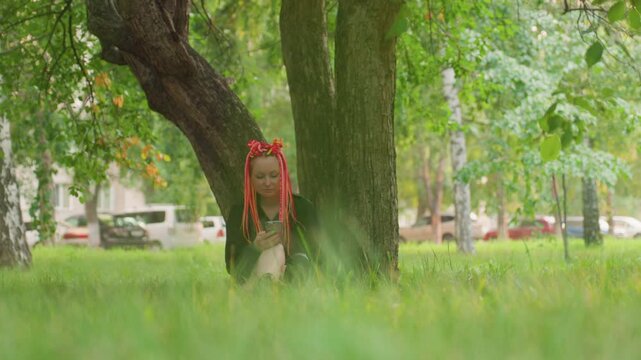 woman relaxes under leafy tree, woman takes peaceful break beneath lush tree in city environment, solitary woman seeks tranquil respite beneath branches amidst bustling city surroundings