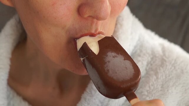 Close-up of a young woman in a bathrobe enjoying a chocolate-coated ice cream bar.