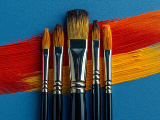 Set of artist paintbrushes with varied bristle shapes and sizes resting on blue canvas with streaks of vibrant red and yellow acrylic paint background