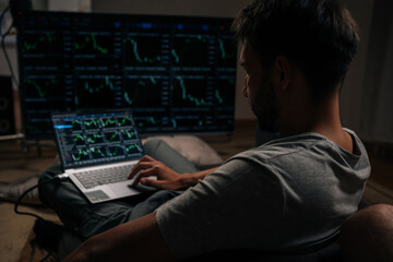 Rear view of successful trader male engrossed in financial analysis and day trading, using laptop and large external monitor displaying various stock market and cryptocurrency charts from home office.